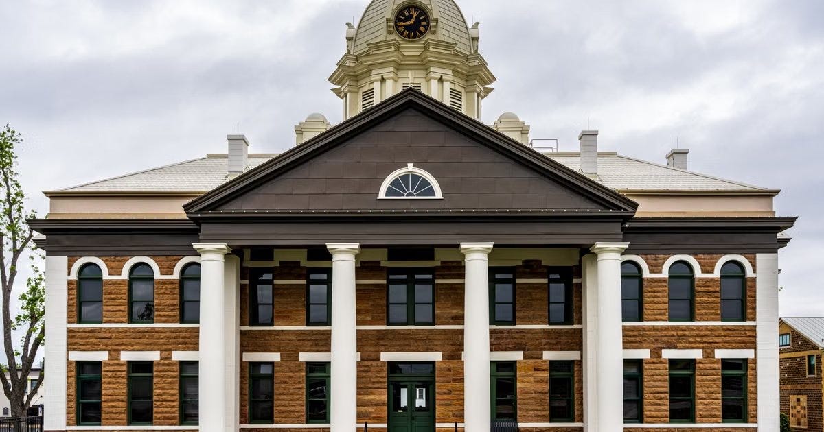 A two-story stone courthouse with four white columns, green-framed arched windows and a clock in the cupola, under an overcast sky