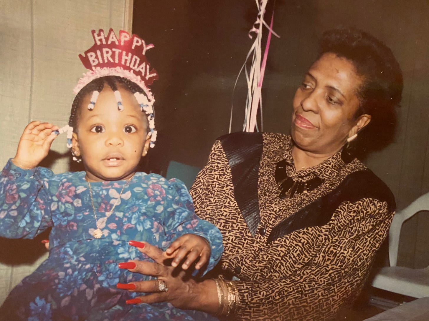 A vintage-style indoor photo of a Black grandmother and a toddler. The grandmother, on the right, wears a patterned blouse and a prominent necklace, looking down and smiling at the child. The toddler, on the left, wears a floral dress and a "Happy Birthday" headband and looks wide-eyed directly at the camera. The grandmother has long, painted red fingernails gently holding the child.