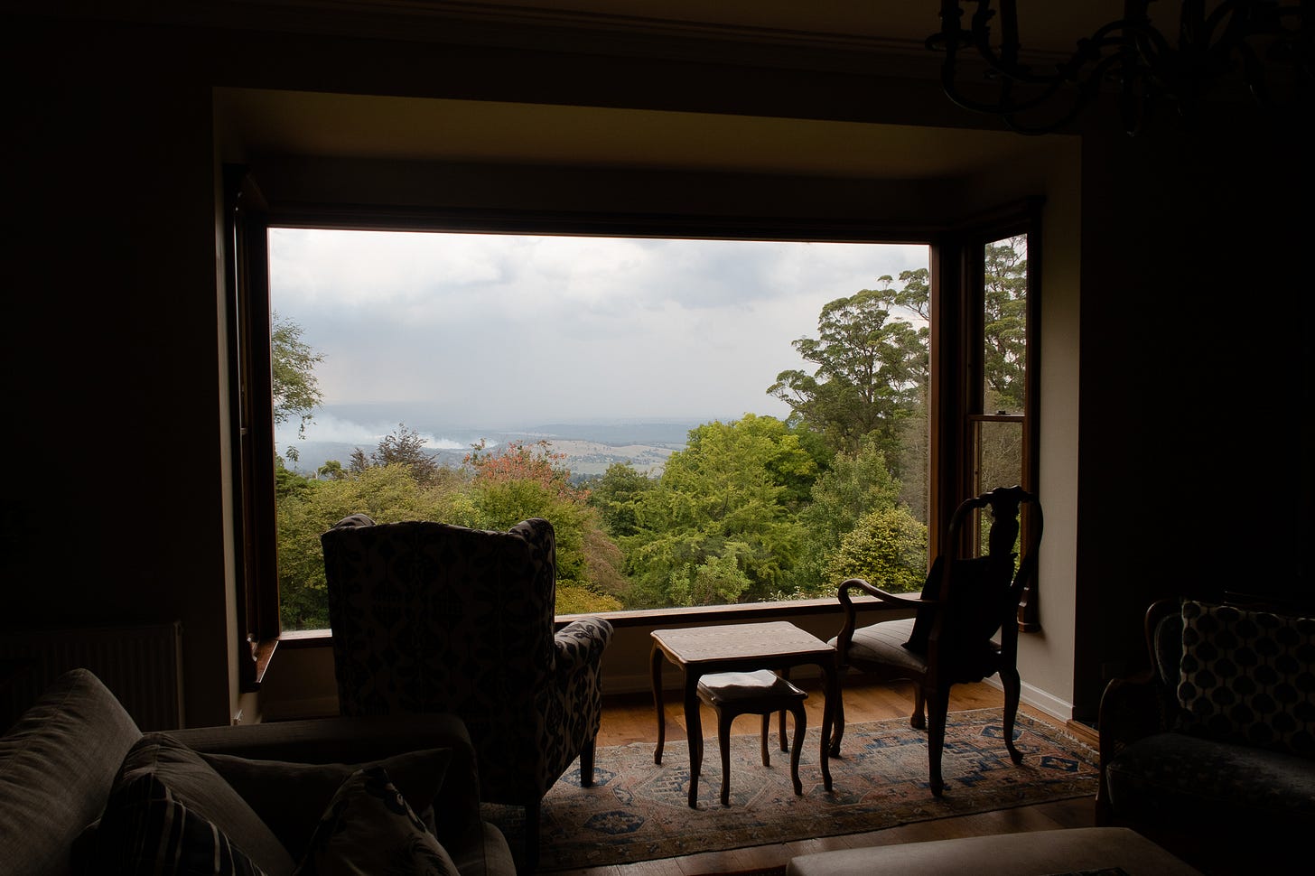 A photograph from inside the living room of a house focused on the view out the big window. The inside of the house is dark and you can see outlines of the old-fashioned furniture and armchairs. The view out the window shows a group of green trees in front of a mountain range and grey sky.
