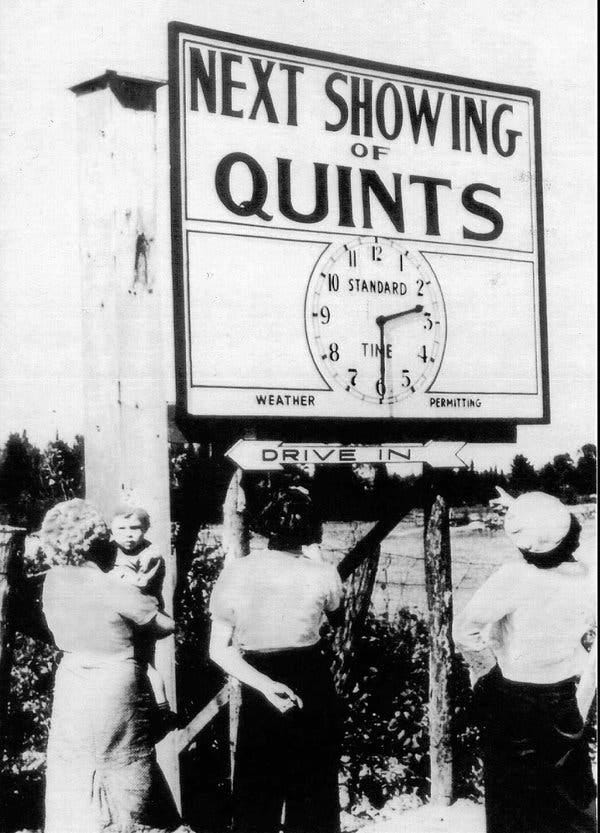 People look up at a sign reading “Next showing of quints” above a clock whose hands indicate 2:30.