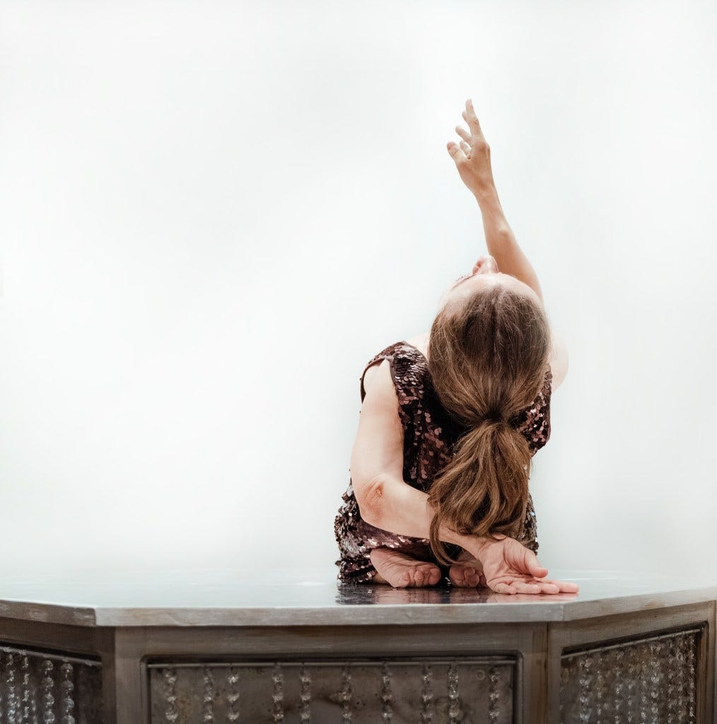 A woman with long brown hair sits cross-legged on a table, one arm behind her back, the other reaching upwards. She wears a sequinned dress and faces away from the camera, set against a plain white background. A woman with long brown hair sits cross-legged on a table, one arm behind her back, the other reaching upwards. She wears a sequinned dress and faces away from the camera, set against a plain white background.