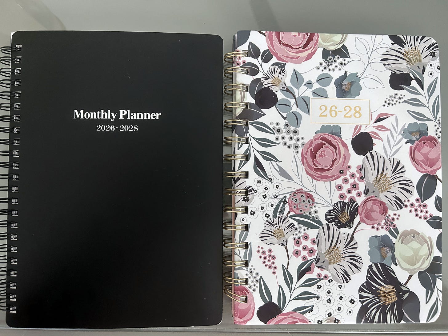A black Monthly Planner 2026-2028 notebook next to a floral-covered three-year planner, standing upright side by side.