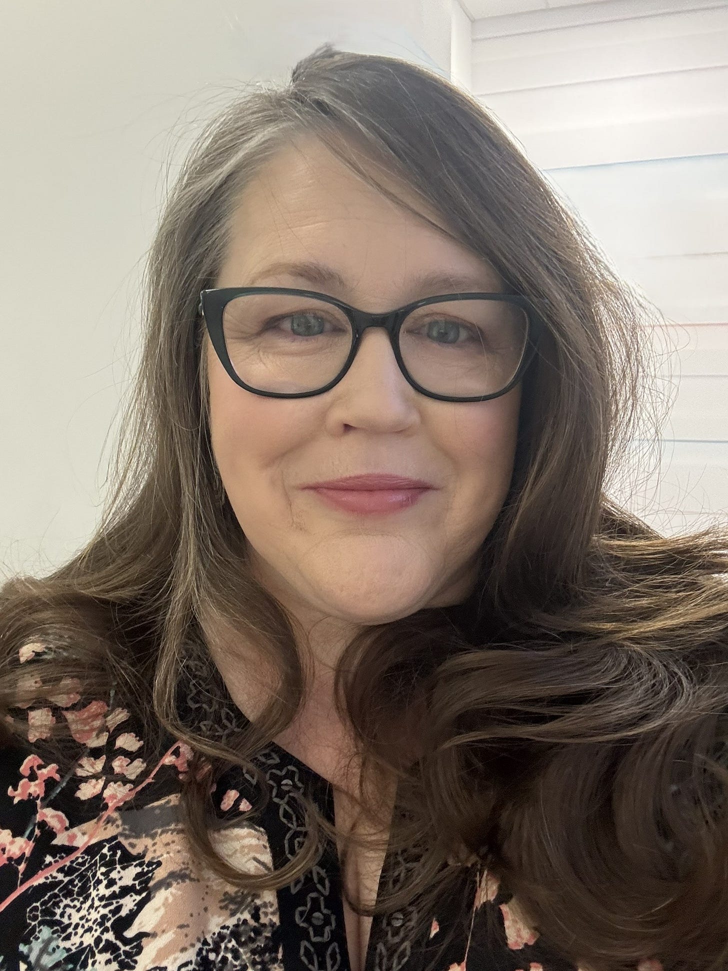 Photo Yolanda Barker,  White woman, long brown hair, middle aged.  plump.  green glasses and a smile.  black dress with orange and white flowers and pattern.