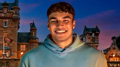 A young man in a light blue hoodie smiles to camera, set against the backdrop of a castle by night A young man in a light blue hoodie smiles to camera, set against the backdrop of a castle by night