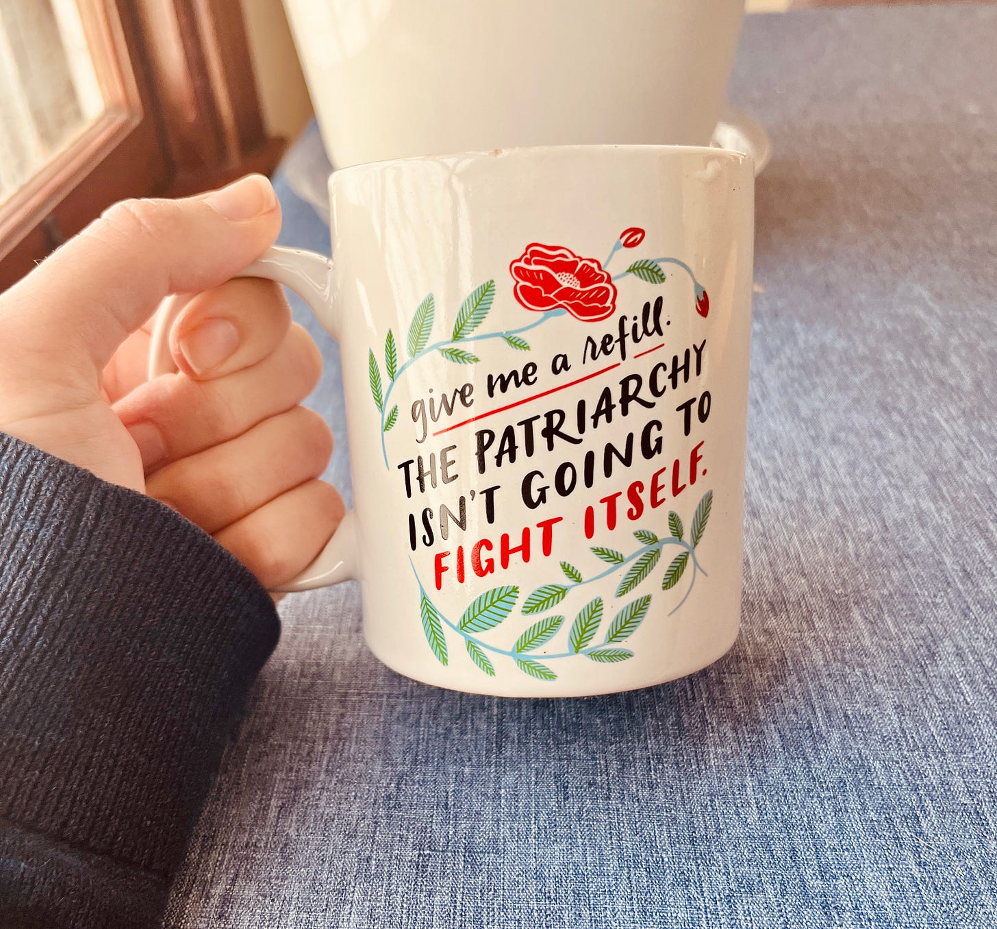 Person holding a coffee mug that reads "Give me a refill. The patriarchy isn't going to fight itself."