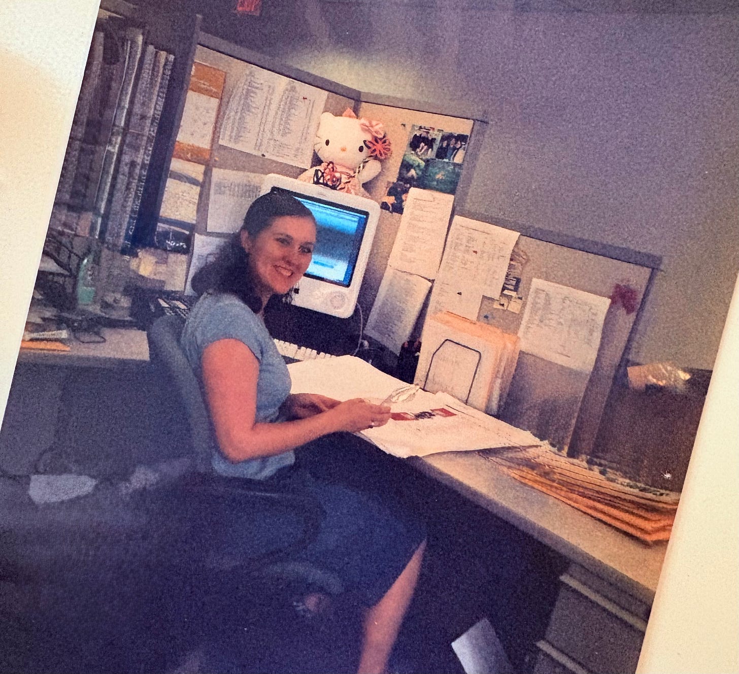 Young Kathryn sits at an open cubicle desk circa 2006/2007, smiling for the camera as she works on magazine layout pages.