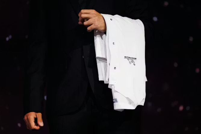 Person wearing black suit holds a white chef's jacket draped over their left arm