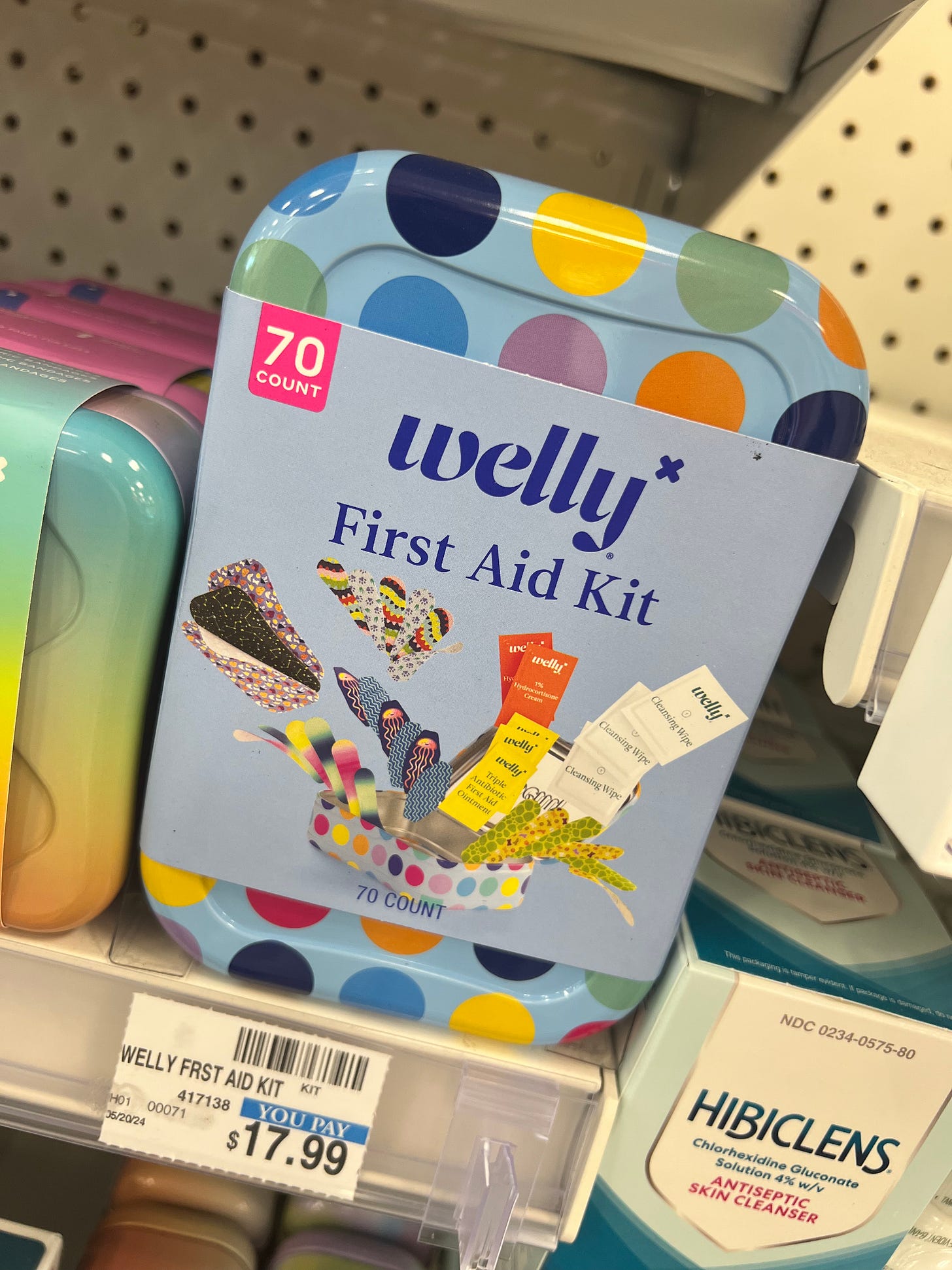 A colorful, polka-dot tin of a Welly First Aid Kit on a store shelf, priced at $17.99. The 70-count kit is shown containing various patterned bandages, cleansing wipes, and triple antibiotic ointment packets. A colorful, polka-dot tin of a Welly First Aid Kit on a store shelf, priced at $17.99. The 70-count kit is shown containing various patterned bandages, cleansing wipes, and triple antibiotic ointment packets.