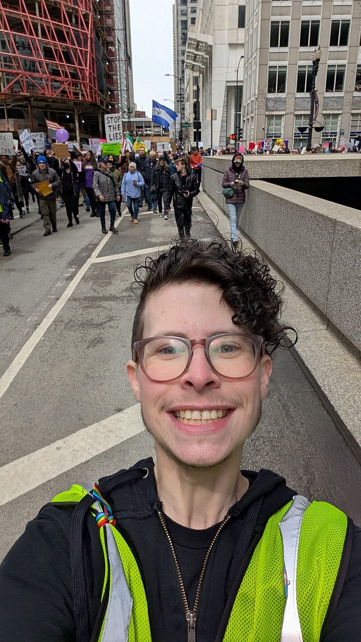 First photo: Asher McMaher (light skin, dark hair, large square glasses, facial hair, black hoodie, black beanie with Trans Up Front logo) marching in protest ahead of long HANDS OFF! banner with large crowd. Second photo: Charlee Friedman (curly hair, large round brown glasses, big smile, facial hair, light skin, wearing a yellow safety vest) ahead of the line of protest marshals and protesters.