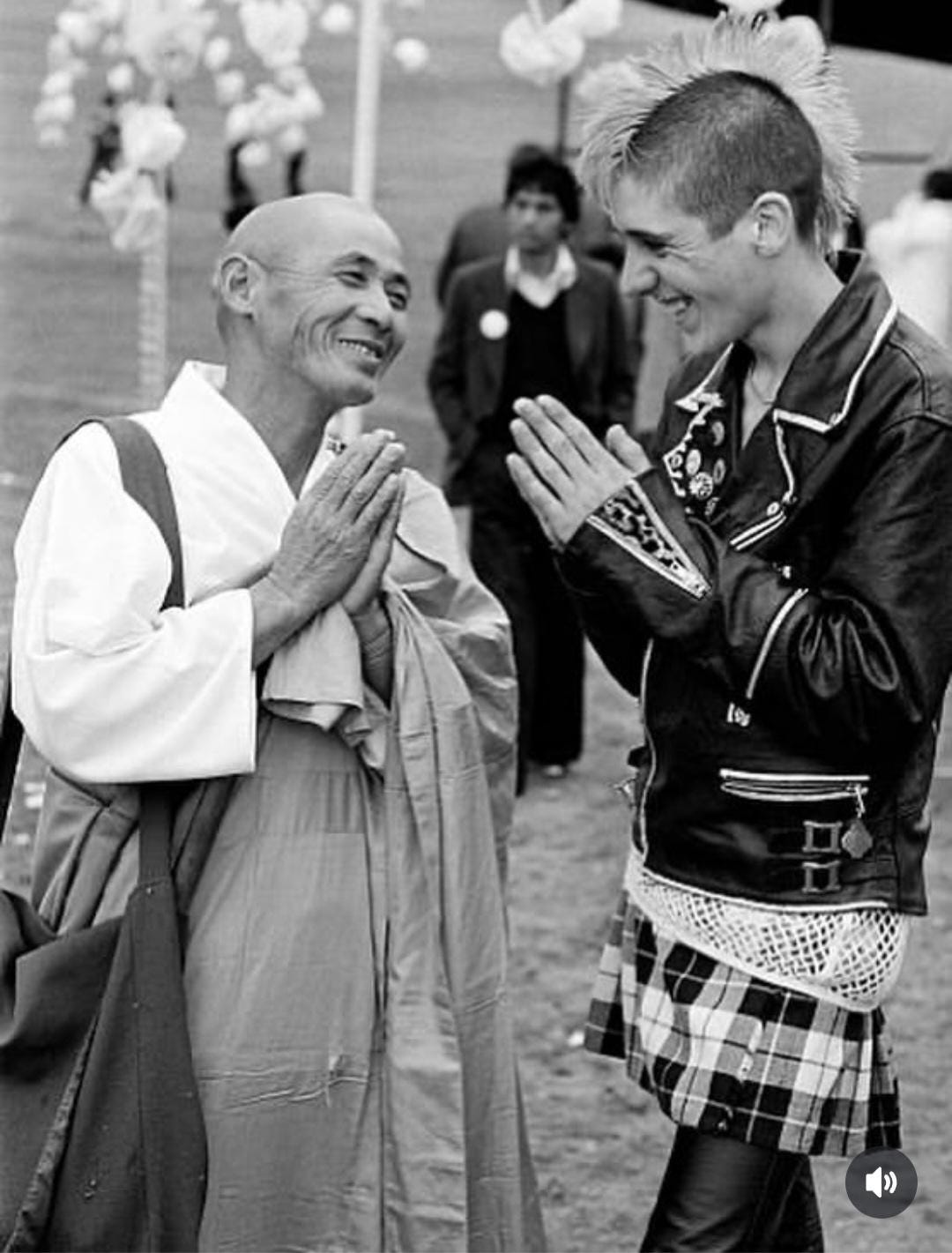 A black and white picture of a monk (left side) and punk (right side). Both have their hands in prayer position and are smiling at each other.
