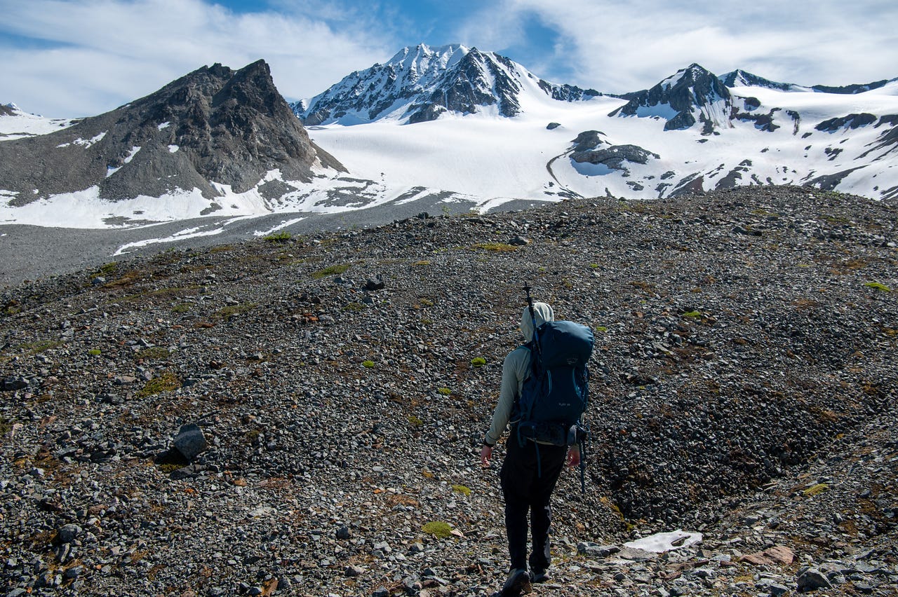 A hiker crosses a broad bench of grey rubble and sparse tundra toward the head of the basin, where a sharp dark rock spire stands to the left of a large snow-plastered peak. Debris-streaked snowfields fill the middle distance under a bright, partly cloudy sky.