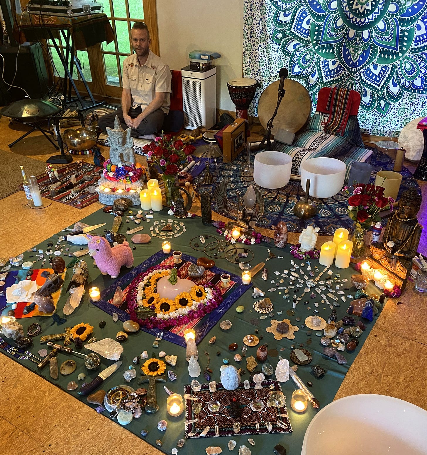 A large crystal grid on the floor surrounded by singing bowls, candles, and sunflowers, with a sound healer seated behind it. A large crystal grid on the floor surrounded by singing bowls, candles, and sunflowers, with a sound healer seated behind it.