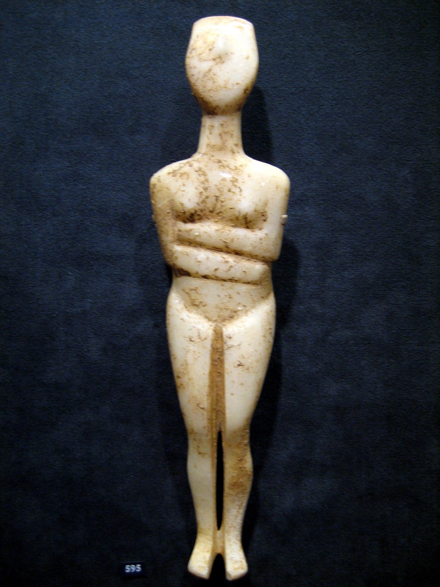White marble Cycladic figurine with brown encrustations on the stone, photographed in front of a dark blue background. The figurine is humanoid, with a schematized body. The only delineated facial figure is a simplified nose. The figure has small breasts. The upper arms are held close against the sides, with the lower arms lying across the upper abdomen just below the breasts, the left arm above the right.