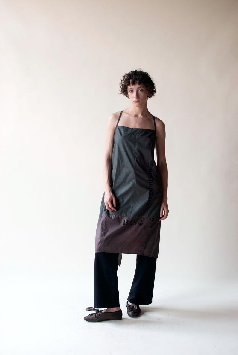 May include: A two-tone apron in shades of gray and brown, with thin straps and a rectangular neckline. The apron is worn over black pants and shoes. The apron has a simple, minimalist design.