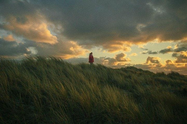 This may contain: a person standing on top of a grass covered hill under a cloudy sky at sunset