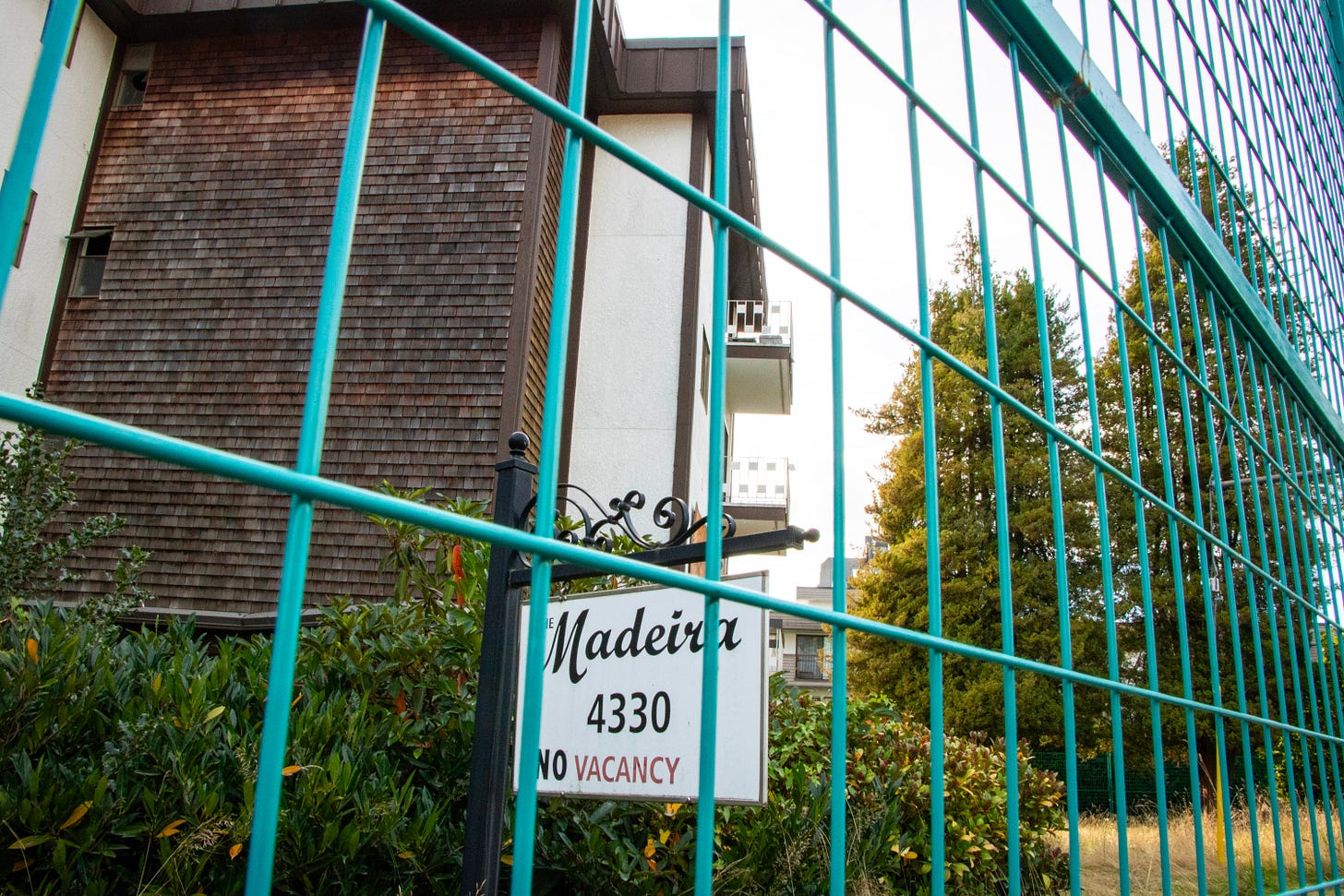 In the immediate foreground, a green metal fence blocks access to a brown and white apartment building, three storeys tall. The sign for the building calls it "The Madeira" and has the number 4330. At the bottom of the sign, it says 'No vacancy'