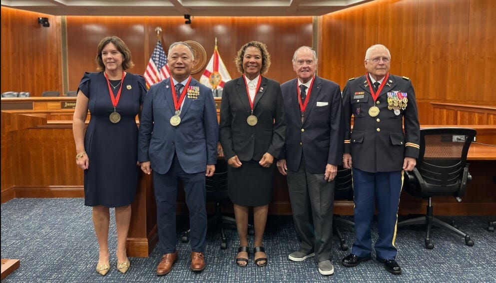 Five adults pose side by side in a wood-paneled courtroom, each wearing a gold medallion on a red ribbon. From left to right: a woman in a navy dress, a man in a blue suit with military decorations, a woman in a black suit, a man in a dark suit, and a man in a formal military uniform with medals. Behind them are desks and chairs, a circular court seal, the United States flag, and two Florida state flags.