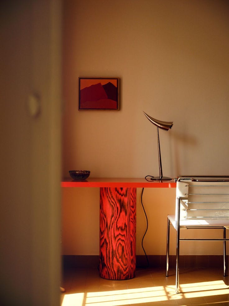 A warmly lit interior corner with a red-orange desk, a small framed artwork on the wall, a sculptural desk lamp, a chair, and sunlight casting shadows across the room. A warmly lit interior corner with a red-orange desk, a small framed artwork on the wall, a sculptural desk lamp, a chair, and sunlight casting shadows across the room.