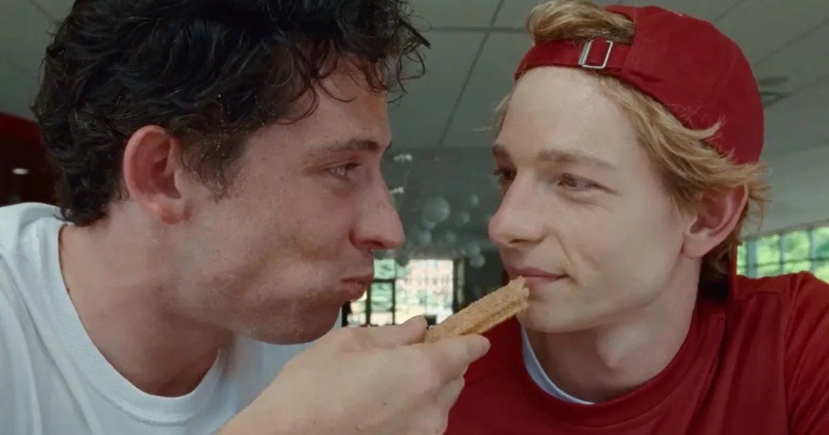 Still from the movie Challengers with a close of up characters Patrick and Art, heads close together, with Patrick holding a churro near Art's mouth