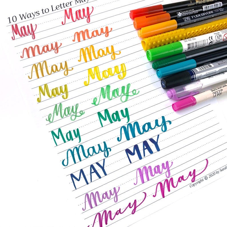 May include: 10 different ways to letter the word "May" in cursive, written in various colors on lined paper. The word "May" is written in pink, red, orange, yellow, green, teal, blue, dark blue, purple, and magenta.  The image also shows a variety of colored brush pens.