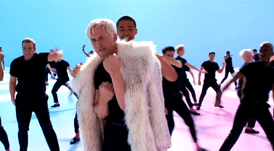 From the I'm Just Ken musical number, Ken approaches the camera with a faux fur draped over his shoulders, holding up his fists in the "put up your dukes" motion. Dozens of other Kens dance behind him in black outfits.