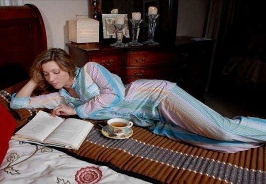 Woman lying on a bed reading a book with a cup of tea beside her.