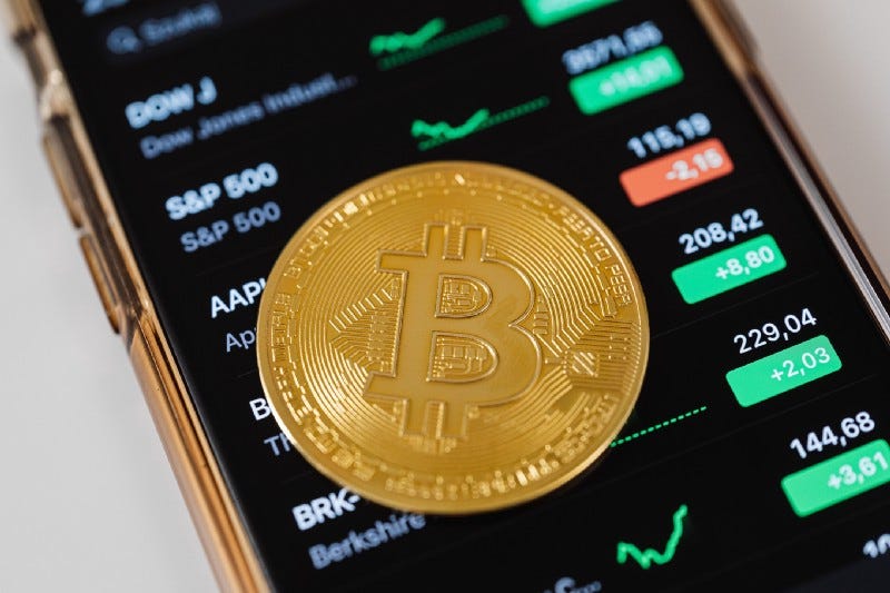 A golden Bitcoin coin on top of a cell phone displaying stock prices. A golden Bitcoin coin on top of a cell phone displaying stock prices.