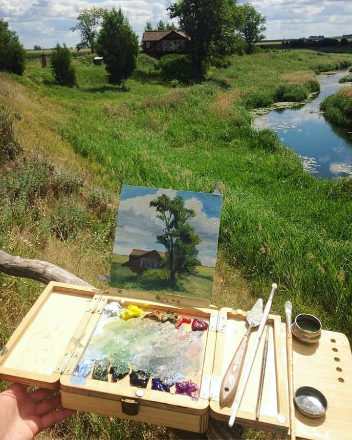 An artist painting a rural landscape with a wooden cabin and tree, using a portable easel box set up by a stream.