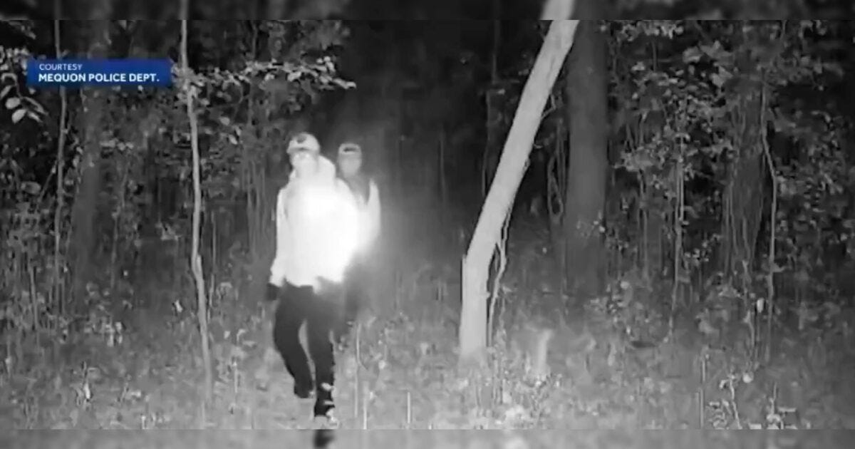 Two individuals walking in a dark forest, illuminated by a flashlight, captured by Mequon Police Department's night vision camera. Two individuals walking in a dark forest, illuminated by a flashlight, captured by Mequon Police Department's night vision camera.