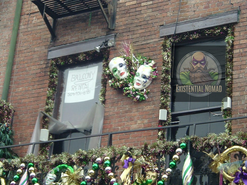 This is a photograph of the exterior of a brick building. The facade is decorated with Mardi Gras garlands, beads, and masks. Two prominent, stylized masks with painted faces are mounted on the wall between two windows. One window has a sign that says "BALCONY RENTAL 504-931-8761," while the other is covered with a black sheet. A metal balcony railing runs along the bottom of the image, also draped with festive beads.