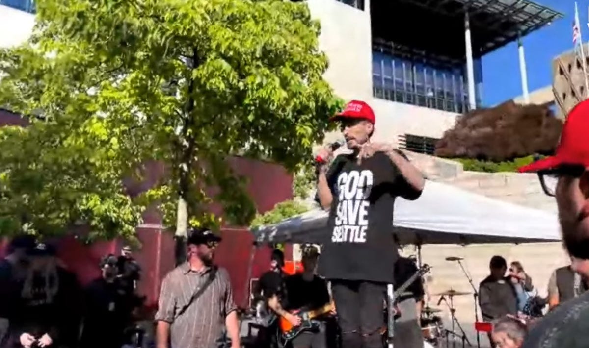 Man wearing a red MAGA-style hat and a T-shirt that says ‘God Save Seattle’ speaks to a small crowd Man wearing a red MAGA-style hat and a T-shirt that says ‘God Save Seattle’ speaks to a small crowd