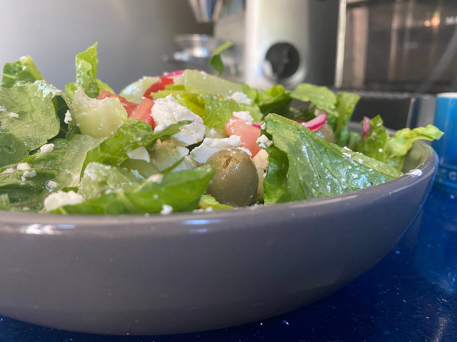 Grey bowl filled with greek cheese, tomatoes, cucumber, lettuce, olives and red onion