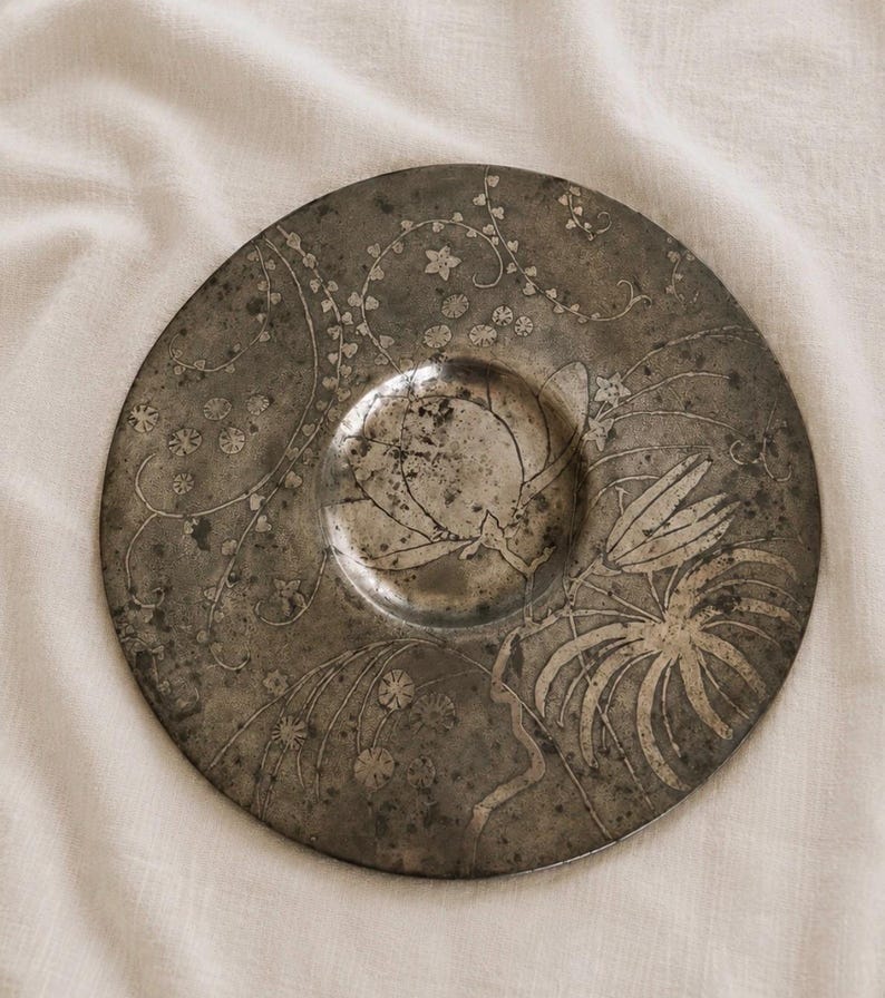 May include: A circular, weathered metal plate featuring a central depression and an etched botanical design. The surface displays a dark, tarnished patina with intricate engravings of trailing vines, small flowers, and large, pointed leaves.