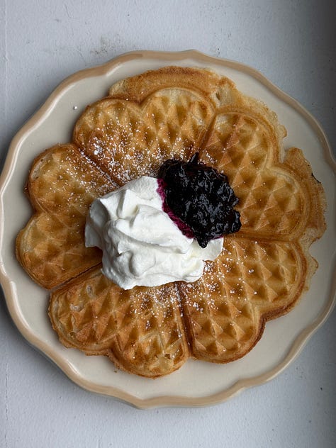 a glass jar with blueberry jam, a swedish waffle with jam and whipped cream, and a wire rack with waffles