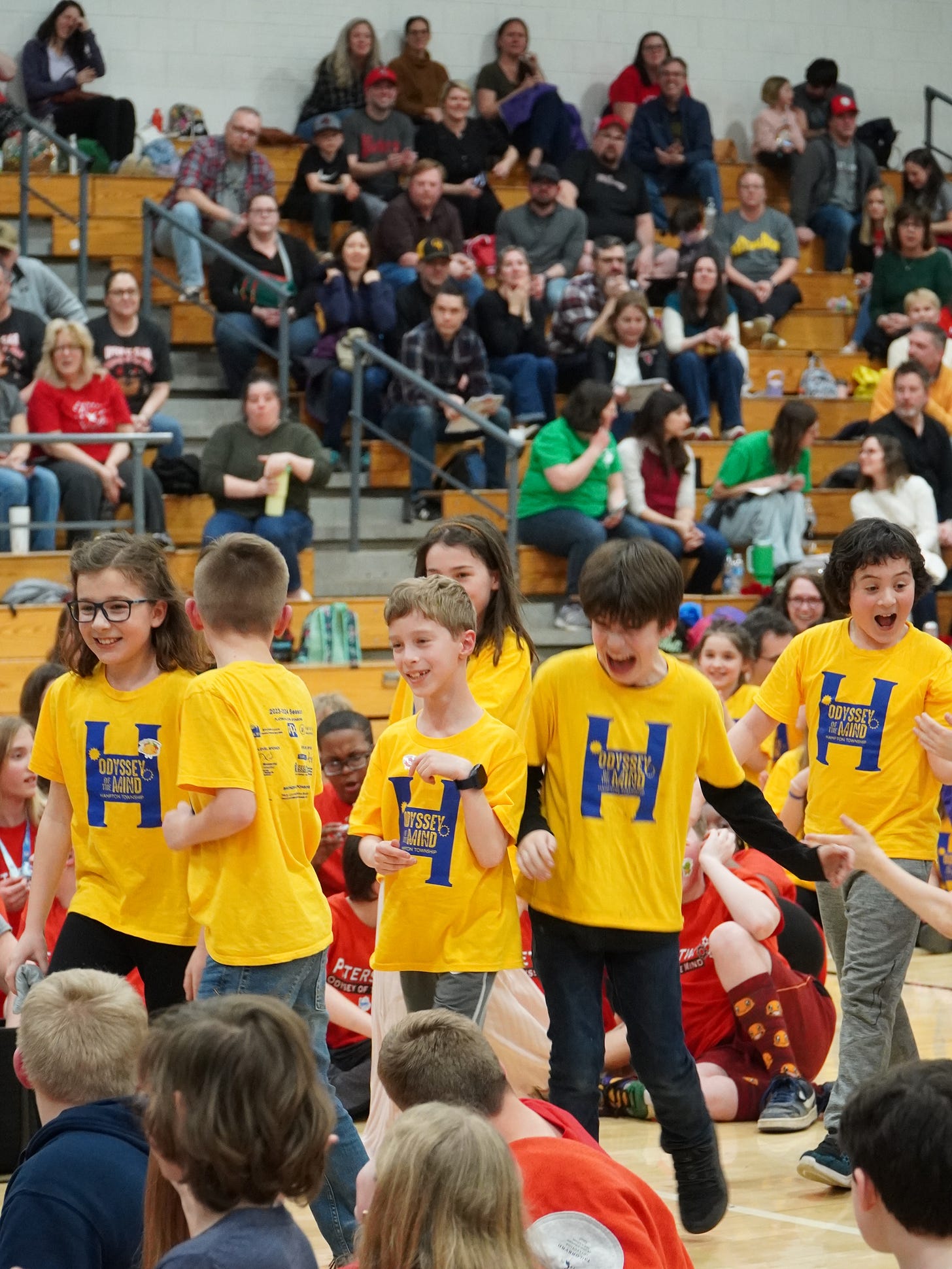 Six children in yellow tee-shirts with Odyssey of the Mind written in blue walk together in a gym. They are smiling, laughing, looking proud. They are surrounded by other children sitting down in different color shirts who are looking at them. Behind him there are adults sitting on the bleachers.