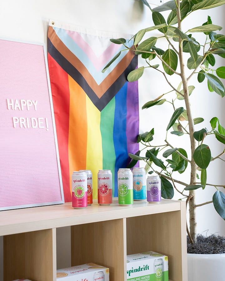 Two images side-by-side, each of six cans of spindrift. In the first, the colors of the cans are, in order, pink, peach, orange, green, blue, turquoise. In the second, the colors are, in order, pink, orange, peach, green, turquoise, purple. The purple can is inexplicably smaller. Behind each there is a progress flag, and the second picture has a letter board that said "happy pride" in white on a light pink background.