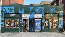 Loyalty's new storefront with an emerald green and light blue mural, a read banned books flag on one door, and Hannah standing in the doorway of the second door. 