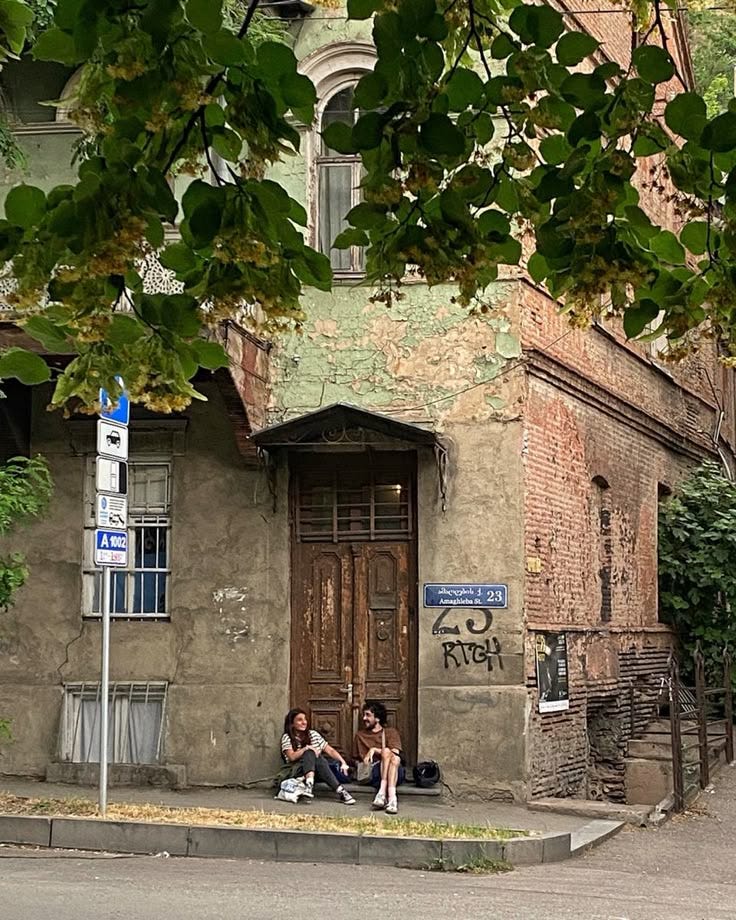 This may contain: two people sitting on the sidewalk in front of an old building with graffiti written on it