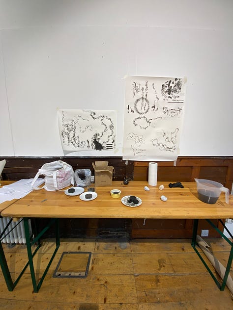 1: a wooden table featuring a selection of tools for mark-making and drawing. 2: a selection of abstract drawings featuring figures and stones in earthy tones. 3: a wooden floor covered in twirling patterns of yellow sand.