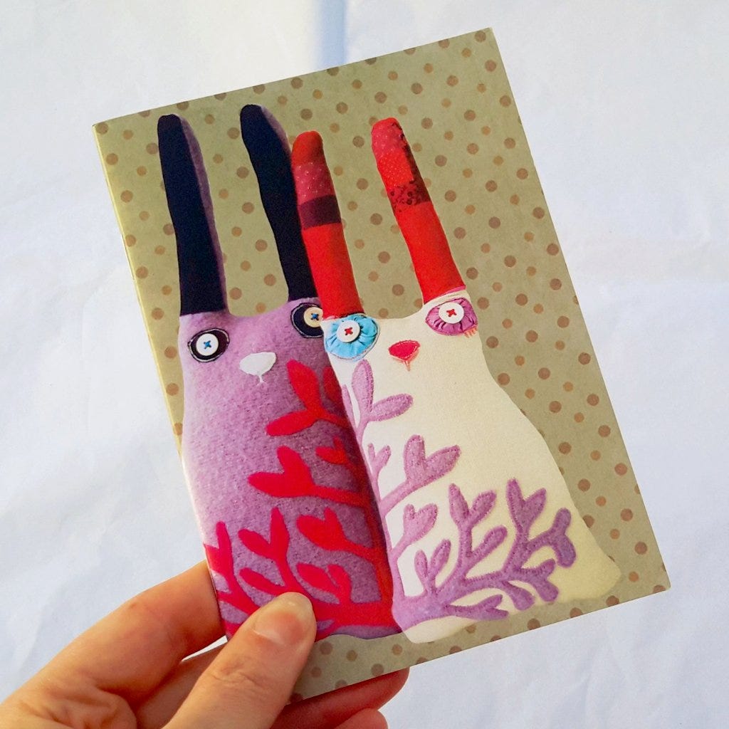 A hand holds up a small notebook with two handmade rabbit dolls on the cover