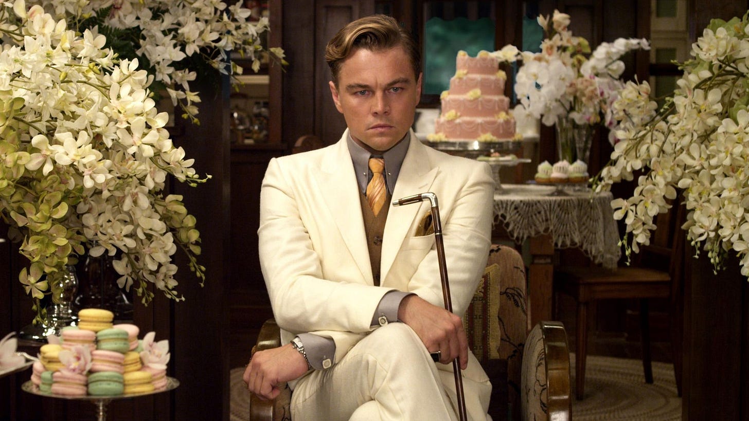 Leonardo DiCaprio as Jay Gatsby. He is wearing a white suit and holding a cane, and appears pensive. He is in a room full of flowers and cakes.