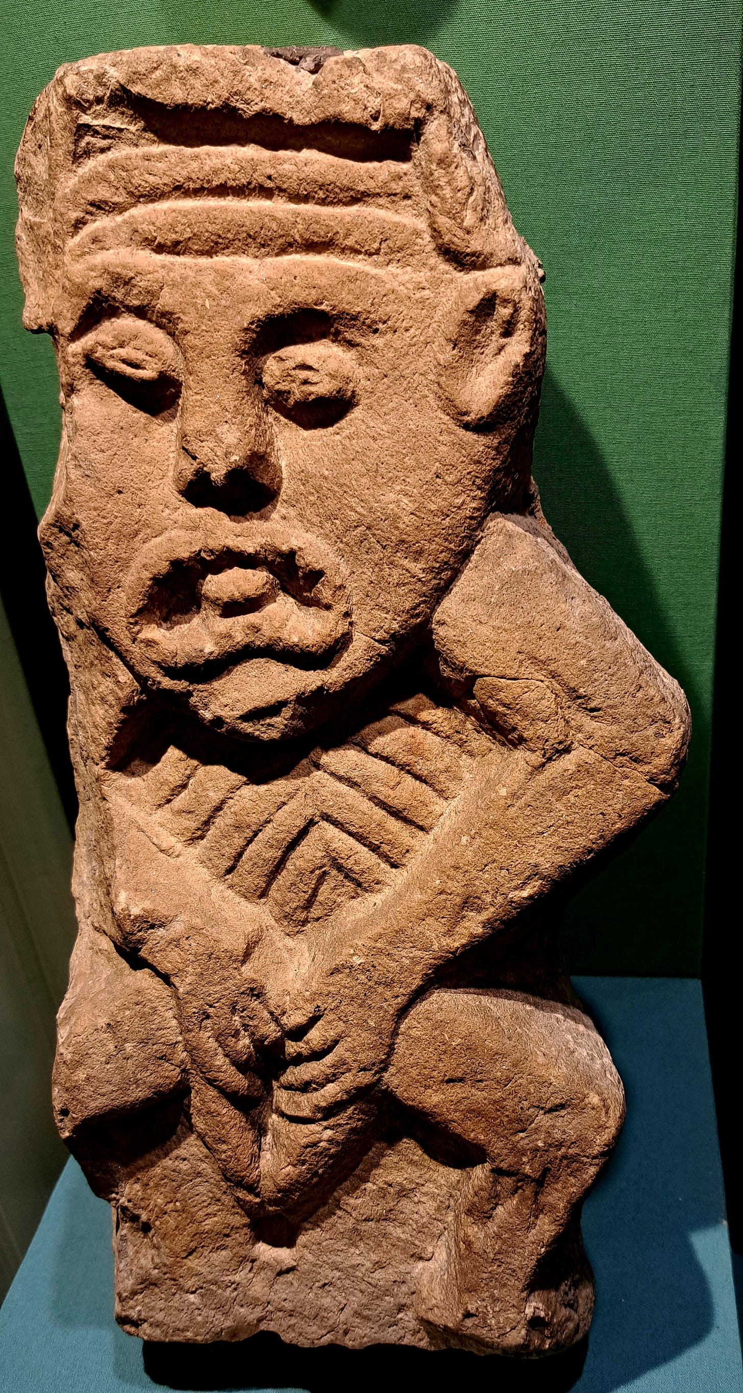 A close-up image of a stone carving of a sheelanagig figure displayed upon a dark green wall. A close-up image of a stone carving of a sheelanagig figure displayed upon a dark green wall.