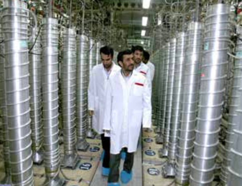 Color photograph of then-President Mahmoud Ahmadinejad tours the Natanz Uranium Enrichment Facility in 2008.  He is accompanied by several men in white lab coats walking through a narrow corridor lined with tall, cylindrical metal centrifuge units on both sides. 