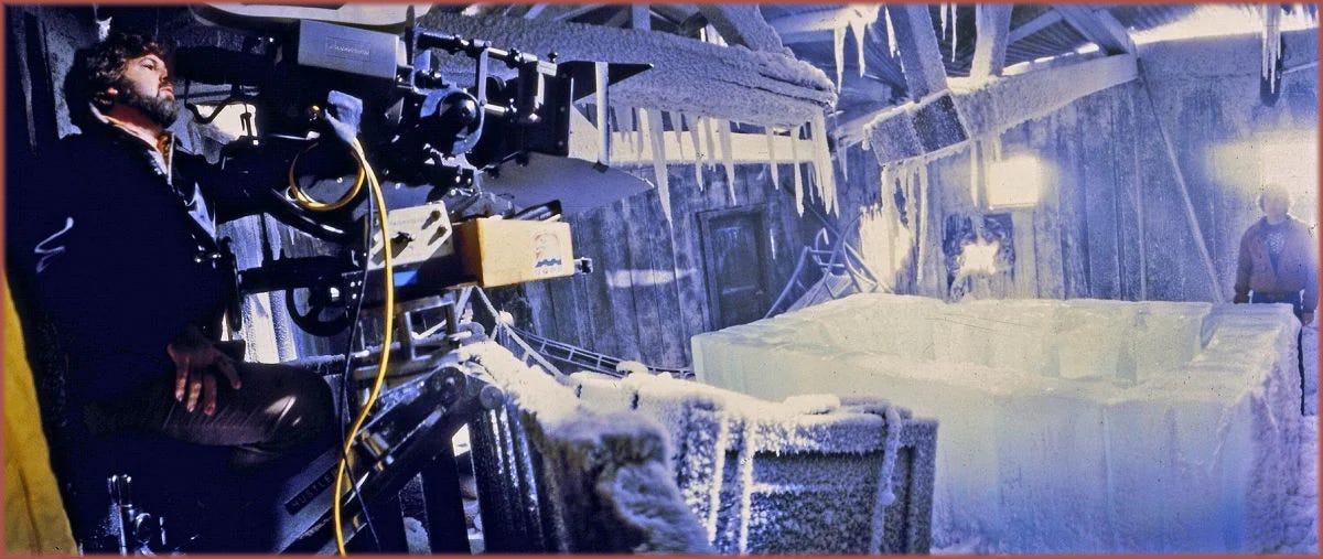 Behind-the-scenes photograph from The Thing (1982): Dean Cundey, ASC crouches beside a Panavision Panaflex camera on the Norwegian camp interior set at Universal Studios, a large block of ice in the foreground and an icy, blue-lit set extending behind him. Photo from the collections of the Margaret Herrick Library.