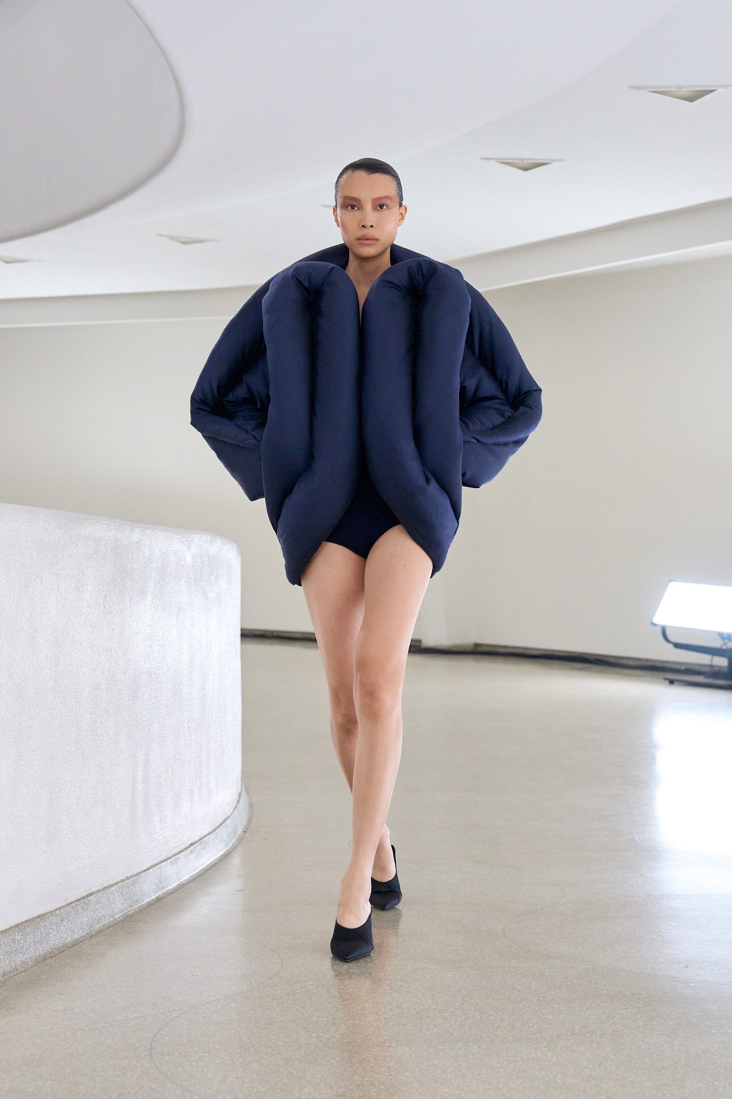 A woman strides down a curved runway while wearing a sculptural, oversized puffer coat, briefs and high heeled shoes.
