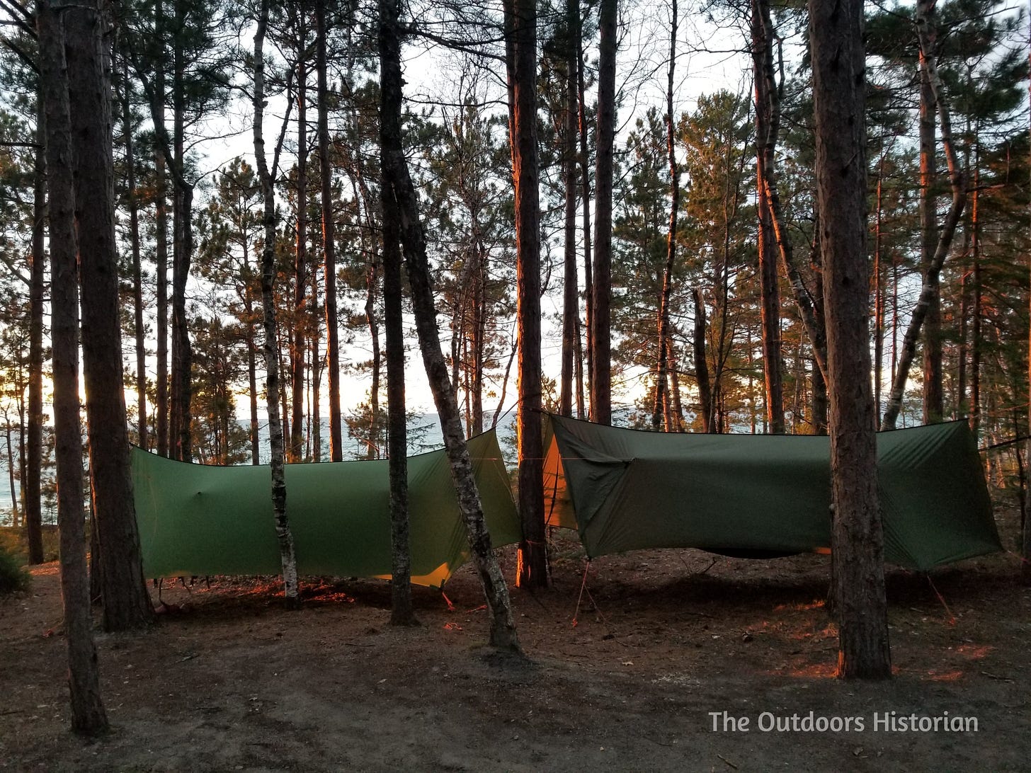 Two hammocks with tarps covering them hang in the pines on a bluff above Lake Superior in the golden glow of a setting sun.