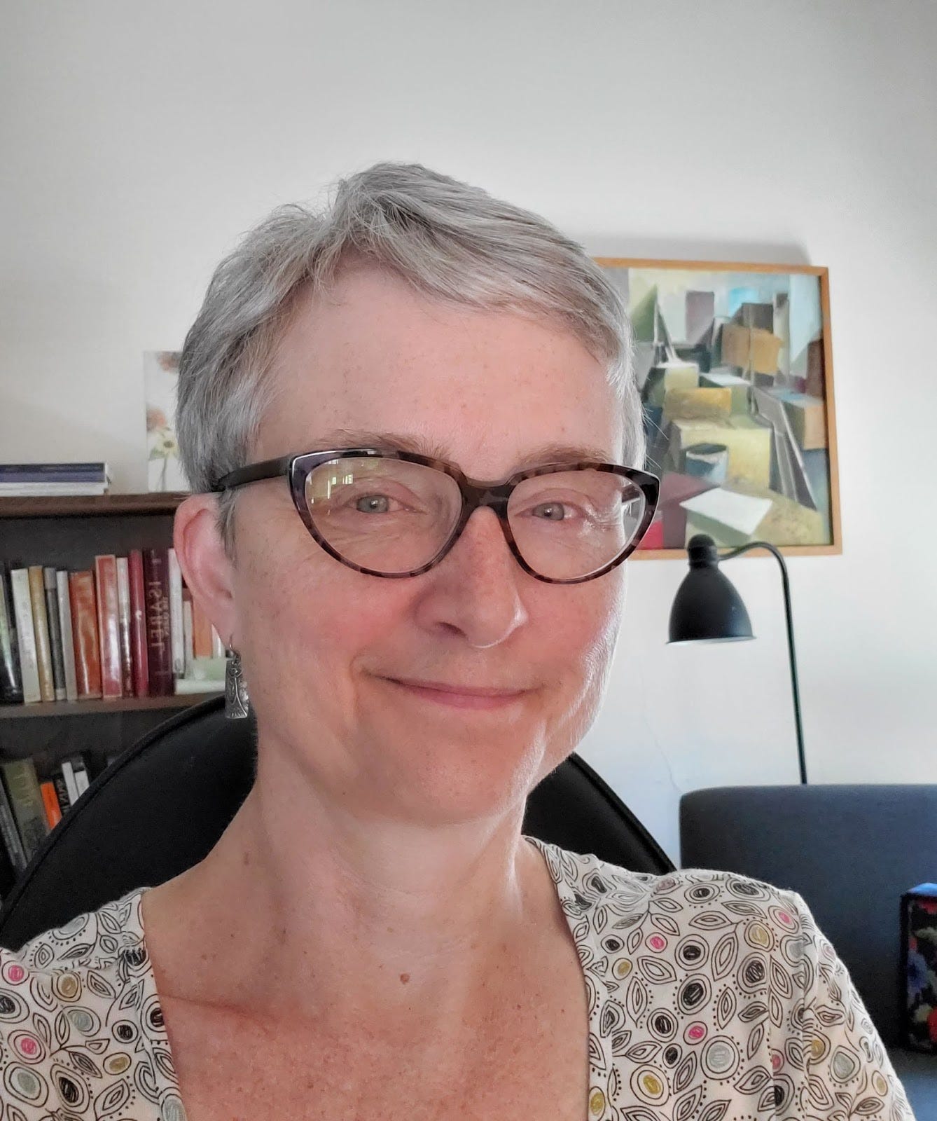 A photo of a white woman with short silve hair, smiling at the camera. She is wearing tortoiseshell glasses, silver earrings, and a cream dress with black and white patterns on it