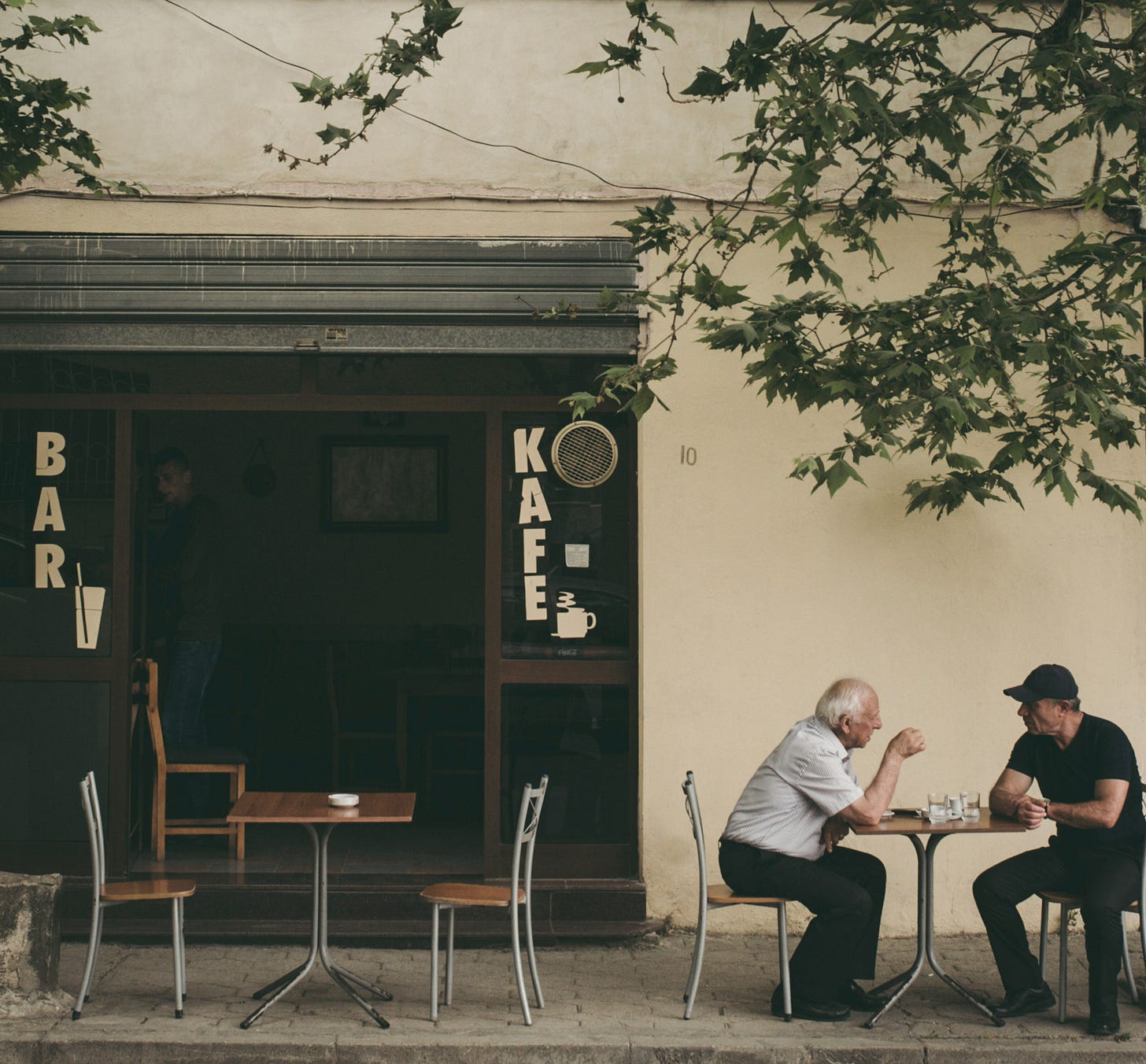 Two elderly men sit at a small round table outside a cafe, engaged in conversation. The cafe has a green storefront with "BAR" and "KAFE" lettering visible on the windows, along with coffee cup symbols. Empty metal chairs and tables are arranged on the sidewalk terrace. Tree branches with green leaves hang overhead, casting dappled shadows on the beige building wall behind them. The scene captures a quiet moment of daily life at what appears to be a European-style neighborhood cafe. Two elderly men sit at a small round table outside a cafe, engaged in conversation. The cafe has a green storefront with "BAR" and "KAFE" lettering visible on the windows, along with coffee cup symbols. Empty metal chairs and tables are arranged on the sidewalk terrace. Tree branches with green leaves hang overhead, casting dappled shadows on the beige building wall behind them. The scene captures a quiet moment of daily life at what appears to be a European-style neighborhood cafe.