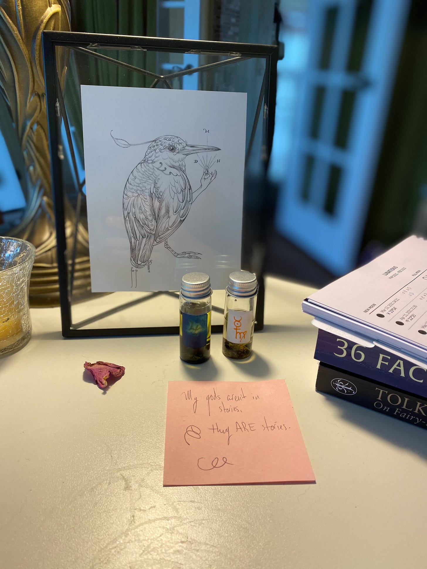 closeup picture of a white desk with a framed image of a bird with a human hand. on the desk there is a stack of books, two small vials of oil, and a pink sticky notes with a handwritten message: "my gods aren't IN stories. my gods ARE stories."
