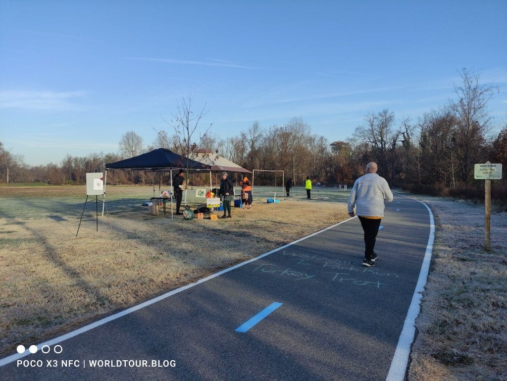 A wide trail is marked with chalk, 'Welcome to parkrun Turkey Trot'. Gazebos are set up on the grass next to the trail for people to leave their bags.